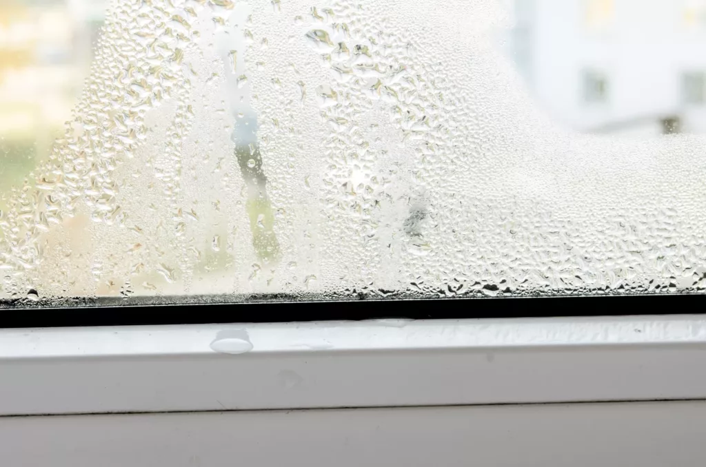 Zaparowana szyba okna z gęstą kondensacją, a krople skraplającej się wody zbierają się u dołu na uszczelce i profilu PVC.