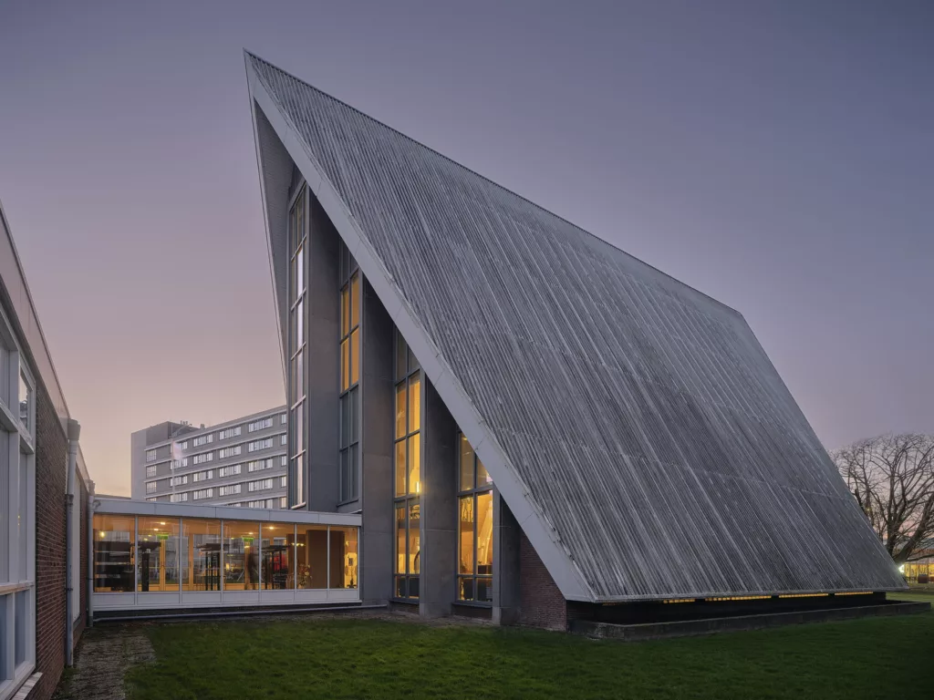 Nowoczesny budynek z bardzo stromym, trójkątnym dachem i wysokimi oknami PVC, oświetlony od środka o zmierzchu; przeszklony łącznik, trawnik na pierwszym planie i blok mieszkalny w tle.