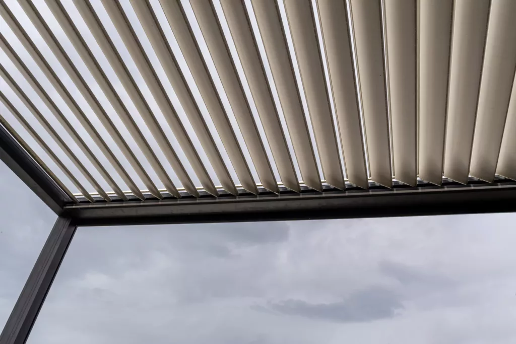 Zadaszenie pergoli z aluminiowymi, ruchomymi lamelami widziane od spodu na tle pochmurnego nieba.