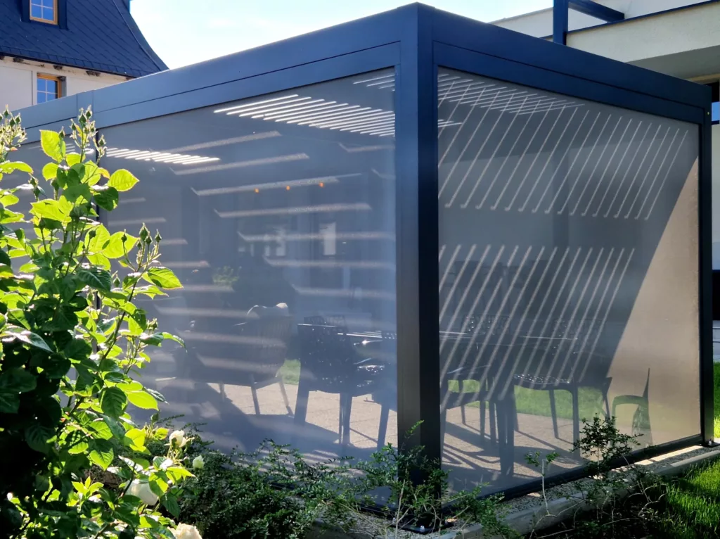 Nowoczesna pergola ogrodowa z ciemną aluminiową ramą i półprzezroczystymi roletami bocznymi, przez które widać cienie lameli oraz zarys stołu i krzeseł, otoczona roślinami przy domu.