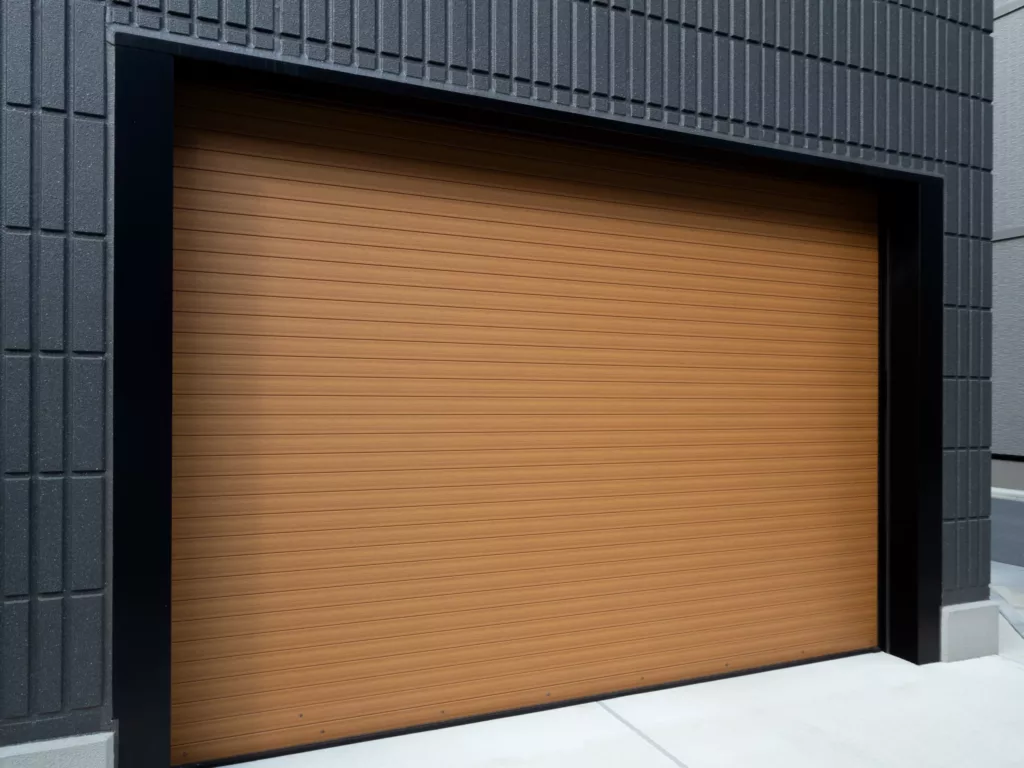 Zamknięta, drewnopodobna brama garażowa rolowana z poziomymi przetłoczeniami w czarnej ramie, osadzona w nowoczesnej, ciemnoszarej elewacji z widocznym betonowym podjazdem.