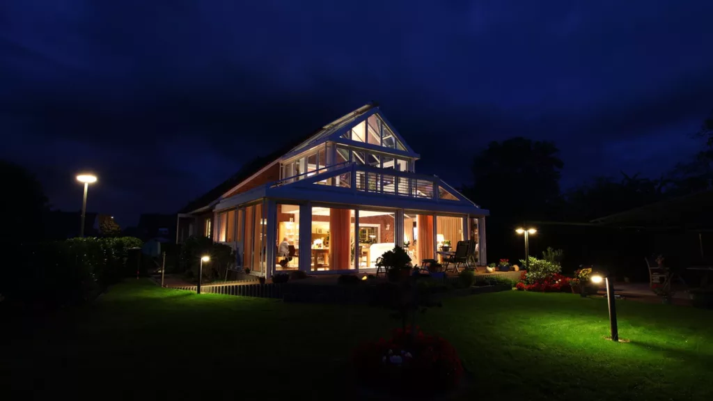Nowoczesny dom z aluminiowym ogrodem zimowych, widziany nocą; ciepłe światło wnętrza i lampy ogrodowe oświetlają taras, zadbany trawnik i rabaty na tle ciemnego, pochmurnego nieba.