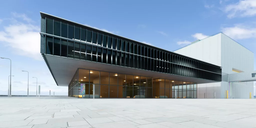 Nowoczesny budynek z dużym, wysuniętym konsolowo piętrem z fasadą aluminiową z ciemnymi żaluzjami, przeszklonym parterem pod postacią fasady aluminiowej i szerokim, pustym placem na pierwszym planie pod błękitnym niebem.