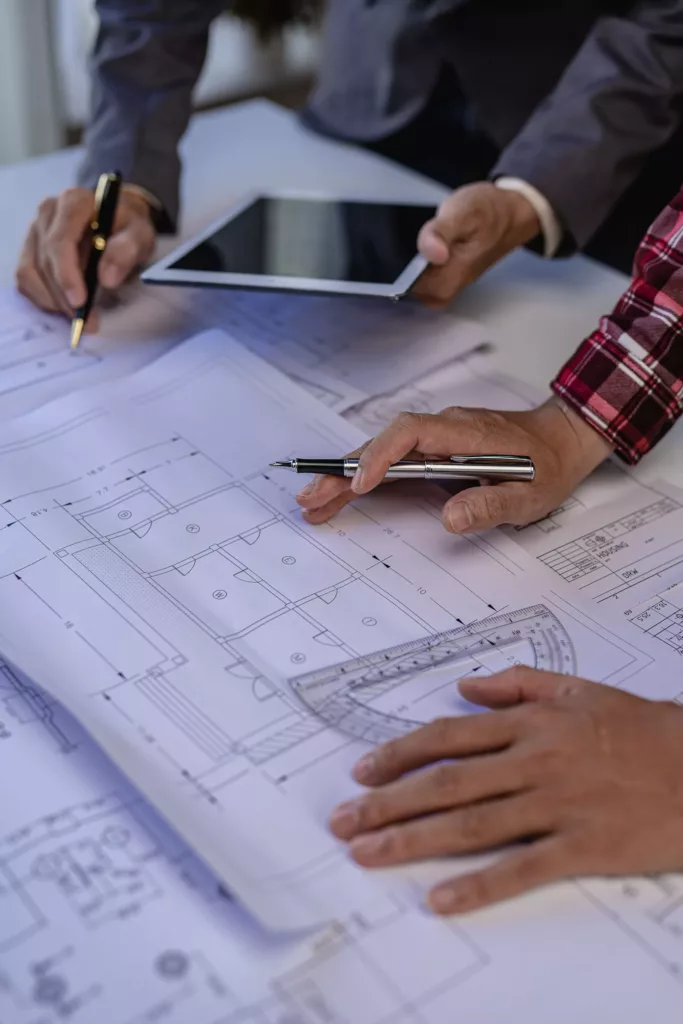 Dłonie dwóch osób nad papierowymi rzutami architektonicznymi, wskazujące długopisami szczegóły projektu obok tabletu i ekierki na biurku.