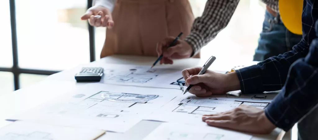 Zespół projektowy omawia na biurku plany architektoniczne, nanosząc uwagi długopisami obok kalkulatora i żółtego kasku ochronnego.