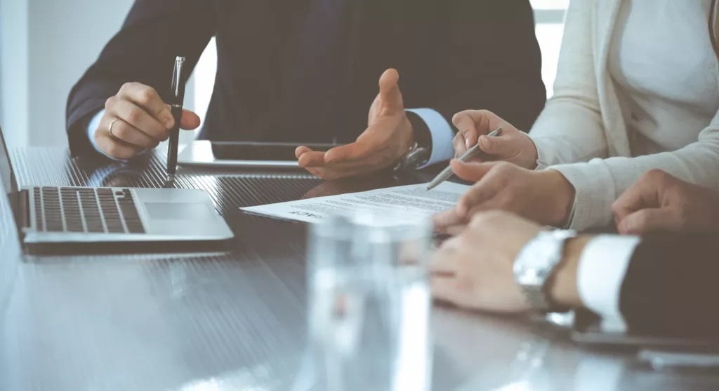 Zbliżenie dłoni osób w strojach biznesowych przy stole konferencyjnym, omawiających projekt i wskazujących na niego długopisami, obok otwartego laptopa.