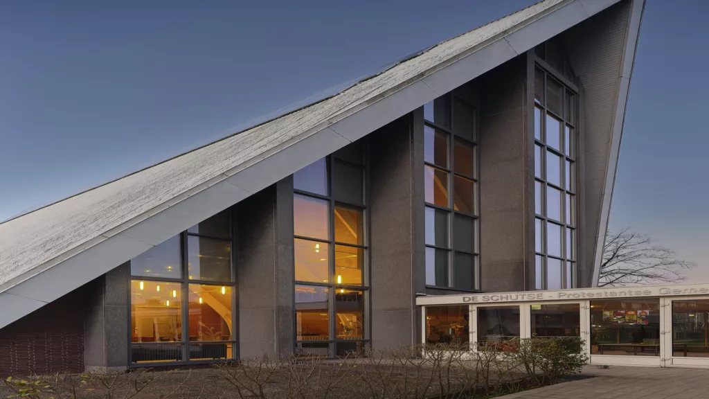 Nowoczesny kościół z bardzo stromym, ukośnym dachem i wysokimi oknam PVCi, sfotografowany o zmierzchu z ciepło oświetlonym wnętrzem oraz napisem „DE SCHUTSE – Protestantse Geme” nad wejściem.