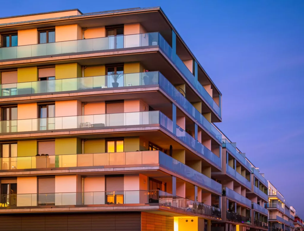 Nowoczesny, wielopiętrowy budynek mieszkalny z szklanymi balkonami, oknami PVC i kolorowymi panelami, odbijający ciepłe światło zachodu na tle niebieskiego wieczornego nieba.