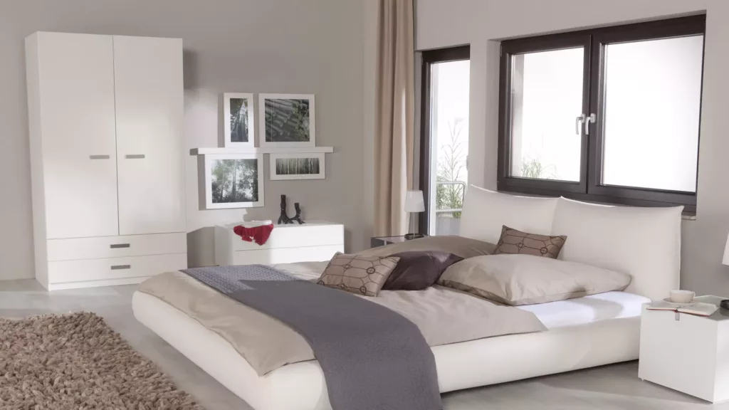 Jasna, nowoczesna sypialnia z niskim białym łóżkiem z beżową pościelą i szarym pledem, dużymi oknami PVC w ciemnych ramach, białą szafą i komodą z grafikami nad nimi oraz miękkim beżowym dywanem i stolikiem nocnym z lampką.