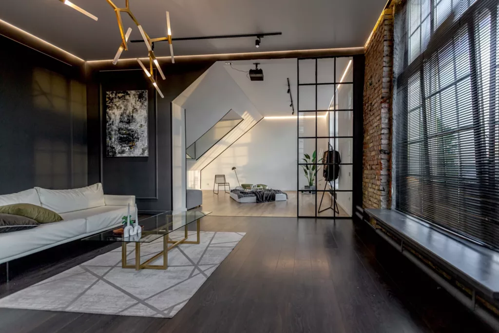 Nowoczesne loftowe wnętrze z ciemną podłogą, białą sofą i szklanym stolikiem na geometrycznym dywanie, dużymi industrialnymi oknami z żaluzjami oraz przeszkloną zabudową wewnętrzną prowadzącą do jasnej strefy sypialnej z niskim łóżkiem i oświetleniem LED.