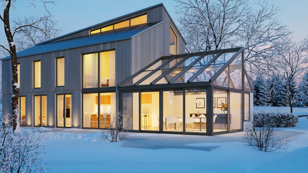 Nowoczesny dom z dużymi oknami i przeszklonym ogrodem zimowym, rozświetlony ciepłym światłem wewnątrz, otoczony zaśnieżonym ogrodem i oszronionymi drzewami o zmierzchu.