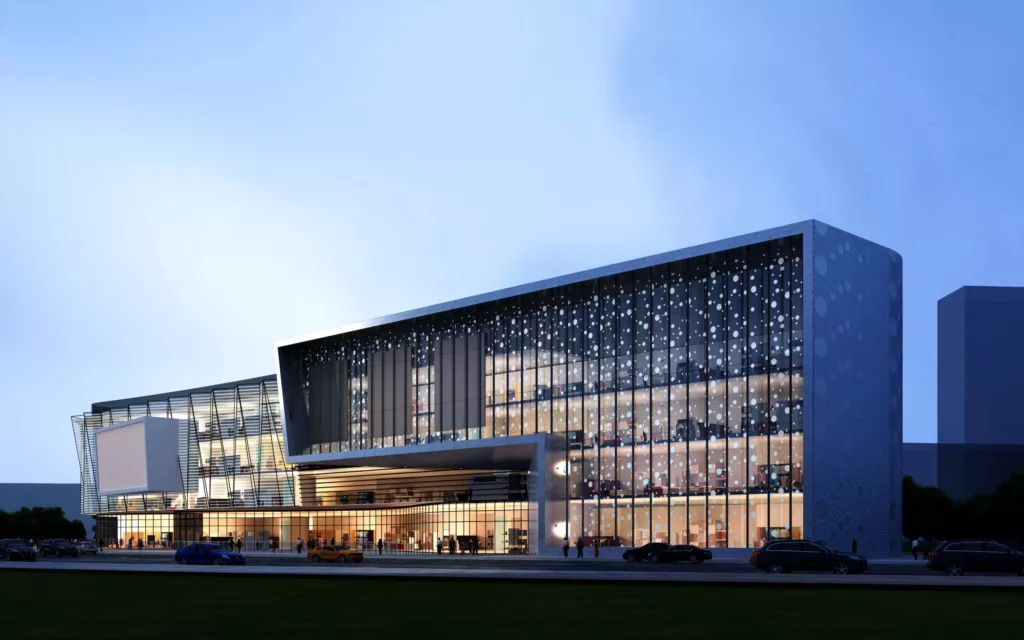 Nowoczesny, wielopoziomowy gmach z dużą przeszkloną fasadą pokrytą wzorem kropek, oświetlony o zmierzchu, z podcieniem wejściowym i samochodami na ulicy przed nim.