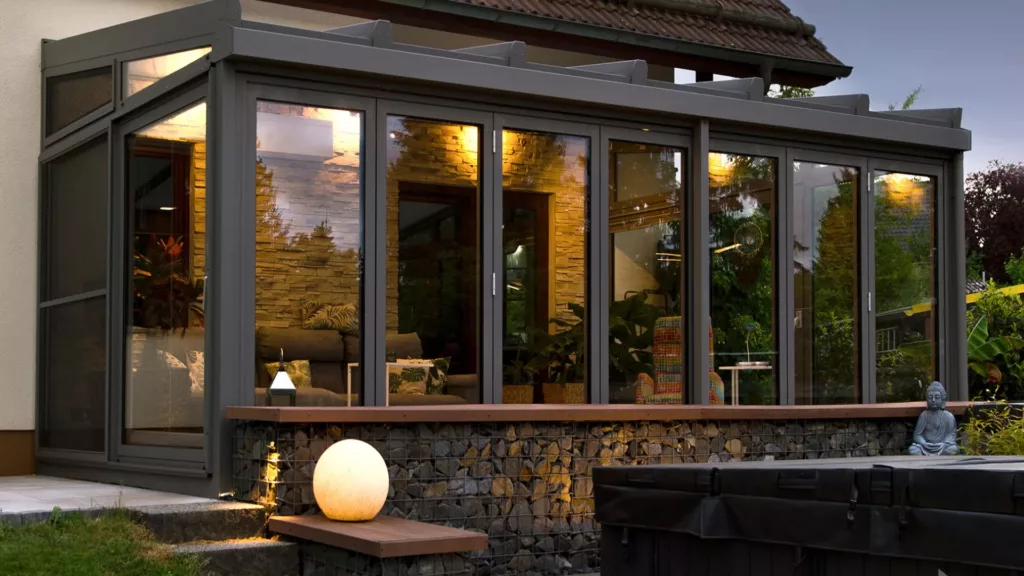 Przeszklona aluminiowy ogród zimowy i przesuwanymi drzwiami, oświetlony ciepłym światłem o zmierzchu; na zewnątrz podmurówka z kulistą lampą i małą figurką Buddy, wokół ogród.