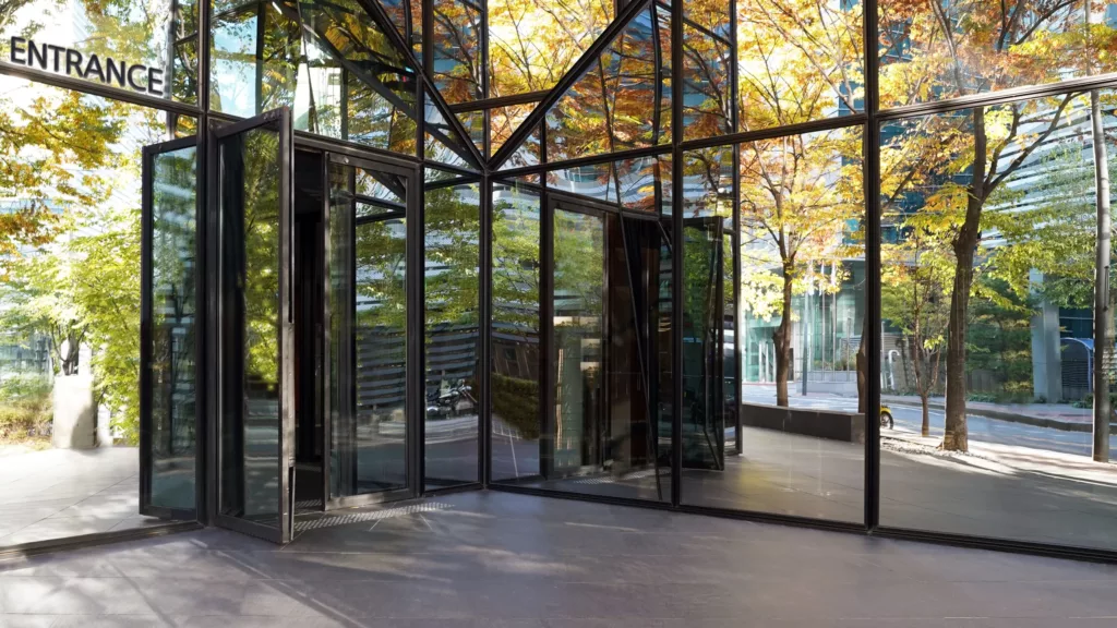 Szklane wejście z obrotowymi drzwiami w nowoczesnym budynku, w którego fasadzie odbijają się jesienne drzewa i ulica.