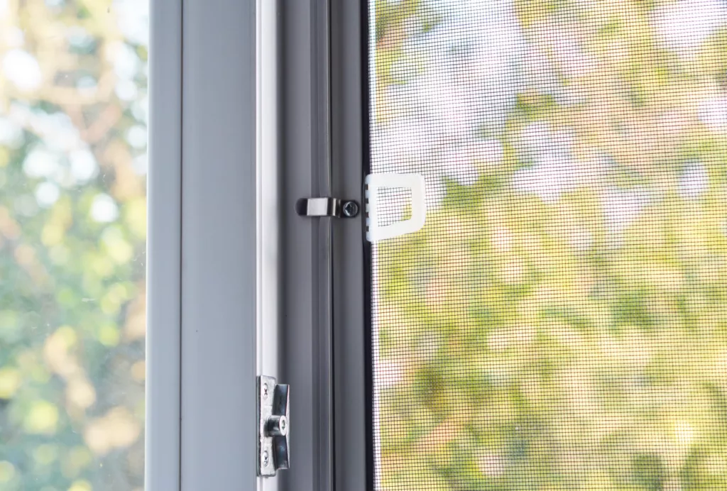 Zbliżenie na ramę okna z zamocowaną moskitierą; widać metalowy zaczep i plastikowy uchwyt siatki na tle rozmytej zieleni za oknem.