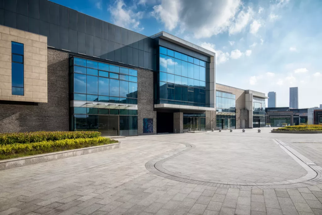 Nowoczesny budynek z fasadą aluminiową z dużymi, lustrzanymi przeszkleniami przy rozległym, pustym, brukowanym placu, z niskimi rabatami i odległymi wieżowcami pod pogodnym, lekko zachmurzonym niebem.
