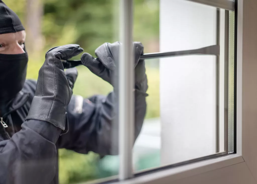 Zamaskowana osoba w czarnych rękawicach używa łomu, aby podważyć okno od zewnątrz.