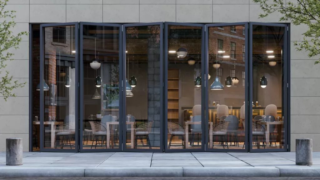 Szklana fasada restauracji z otwartymi, składanymi drzwiami harmonijkowymi; wewnątrz widać stoliki, krzesła i wiszące lampy, a w szybach odbija się elewacja sąsiedniego budynku.