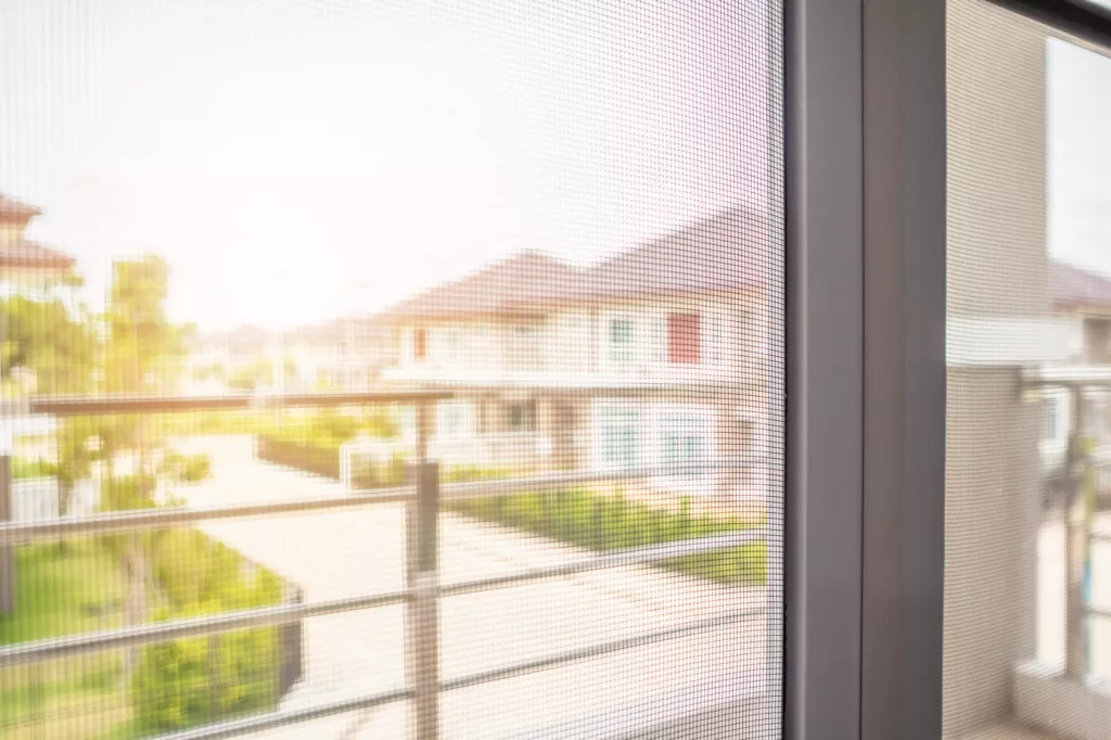 Widok przez siatkę moskitiery w drzwiach balkonowych na słoneczne osiedle, z balustradą na pierwszym planie i rozmytymi domami w tle.