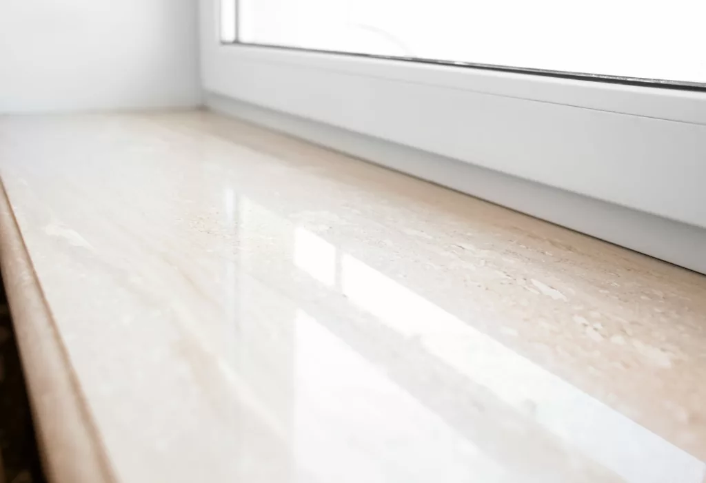 Polerowany beżowy parapet granitowy pod białą ramą okienną z PVC, z wyraźnym połyskiem i odbiciem światła.