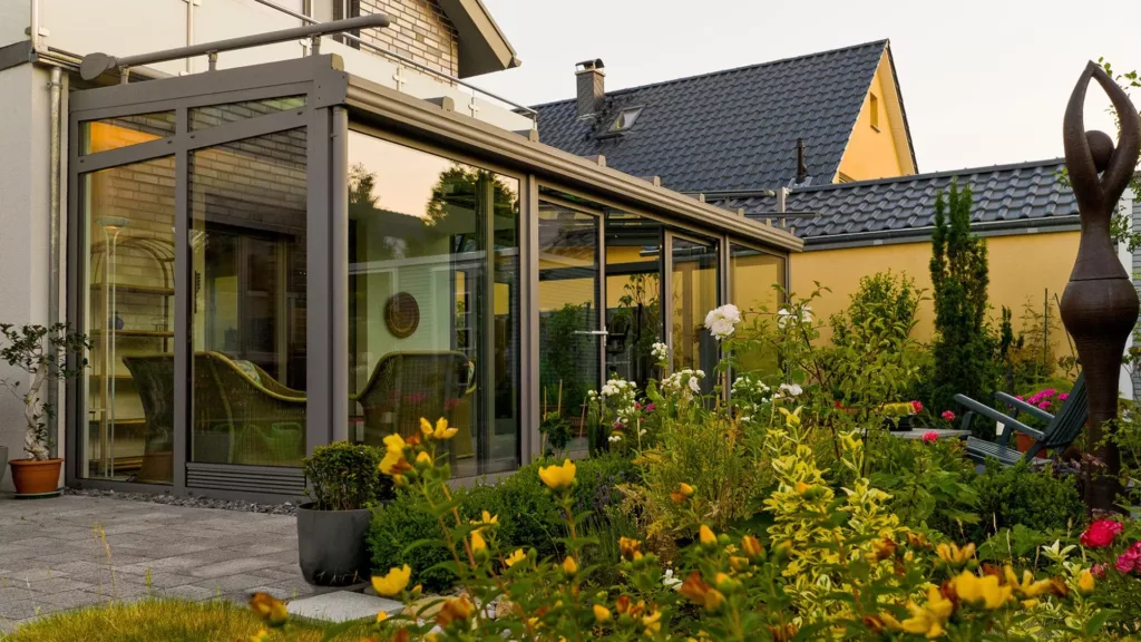 Przeszklony ogród zimowy z aluminium przy domu jednorodzinnym, otoczony bujnymi kwiatami i krzewami, z rzeźbą.
