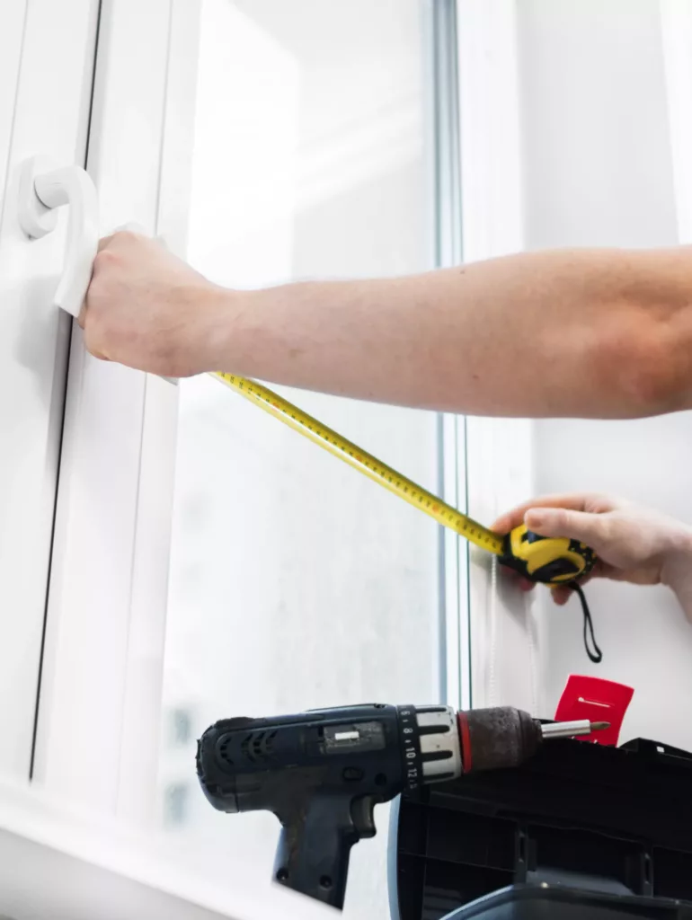 Ręce mierzą taśmą mierniczą szerokość białego okna, a obok na parapecie leży wiertarka i skrzynka narzędziowa.