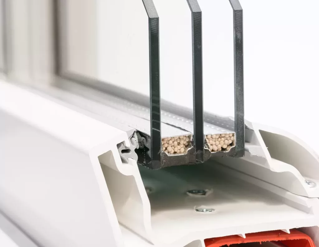 Przekrój okna PVC z pakietem trzyszybowym, widocznymi przekładkami dystansowymi wypełnionymi pochłaniaczem wilgoci oraz elementami ramy (uszczelki, wkręty).