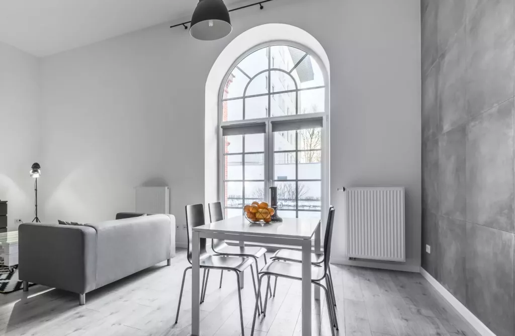 Jasny, minimalistyczny salon z jadalnią: duże łukowe okno ze szprosami, biało‑szara kolorystyka, stół z czterema krzesłami i misą pomarańczy, szara kanapa oraz betonowa ściana akcentowa.