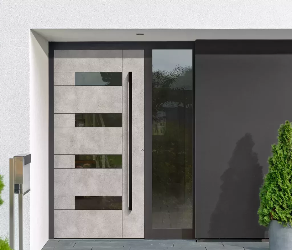 Nowoczesne drzwi wejściowe w szarym, betonowym wykończeniu z poziomymi panelami i wąskimi przeszkleniami, wysoką pionową klamką oraz przylegającym pełnowysokim naświetlem po prawej.