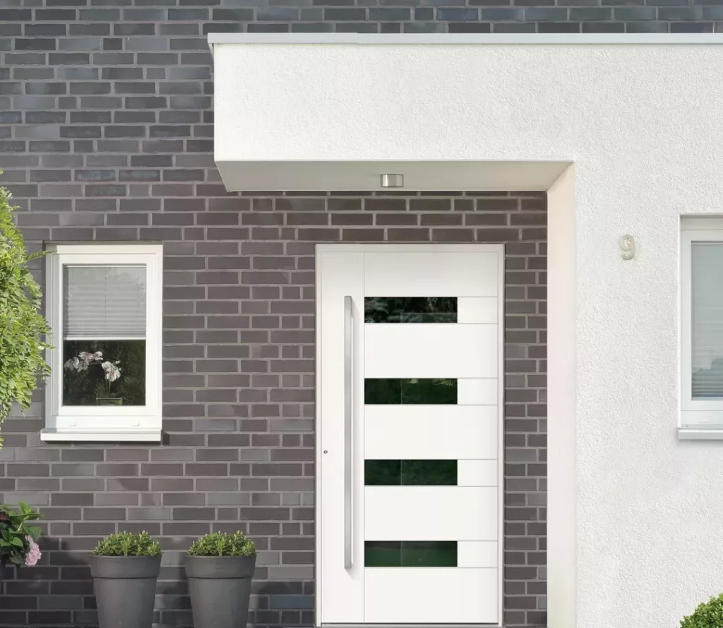 Nowoczesne białe drzwi wejściowe z długim pionowym uchwytem i poziomymi pasami szkła w szarej ceglanej elewacji, pod prostym zadaszeniem, obok okna i donic z roślinami.