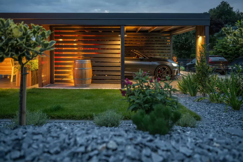 Nowoczesna wiata garażowa z ciepłym oświetleniem, pod którą stoi ciemny kabriolet; obok ściana z poziomych desek i dekoracyjna drewniana beczka, na pierwszym planie zadbany ogród z żwirem i roślinami o zmierzchu.