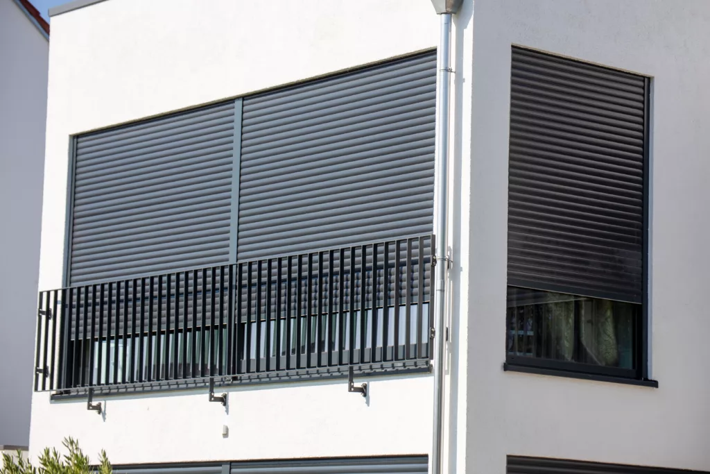 Nowoczesna elewacja w białym tynku z metalową balustradą balkonu i zewnętrznymi, czarnymi roletami zewnętrznymi na dużych oknach, z których jedna jest częściowo opuszczona.
