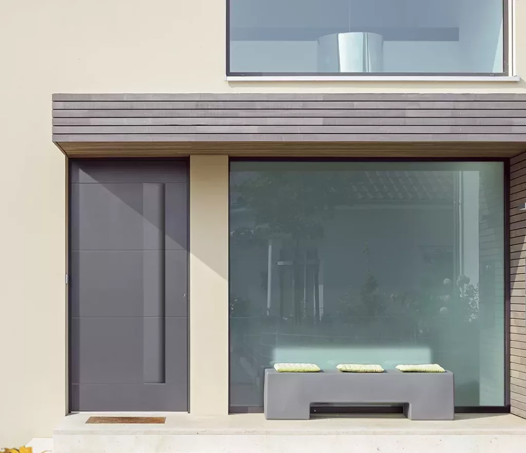Minimalistyczna fasada domu: grafitowe drzwi wejściowe obok dużego, matowego okna oraz szara ławka z trzema jasnymi poduszkami pod poziomym zadaszeniem.