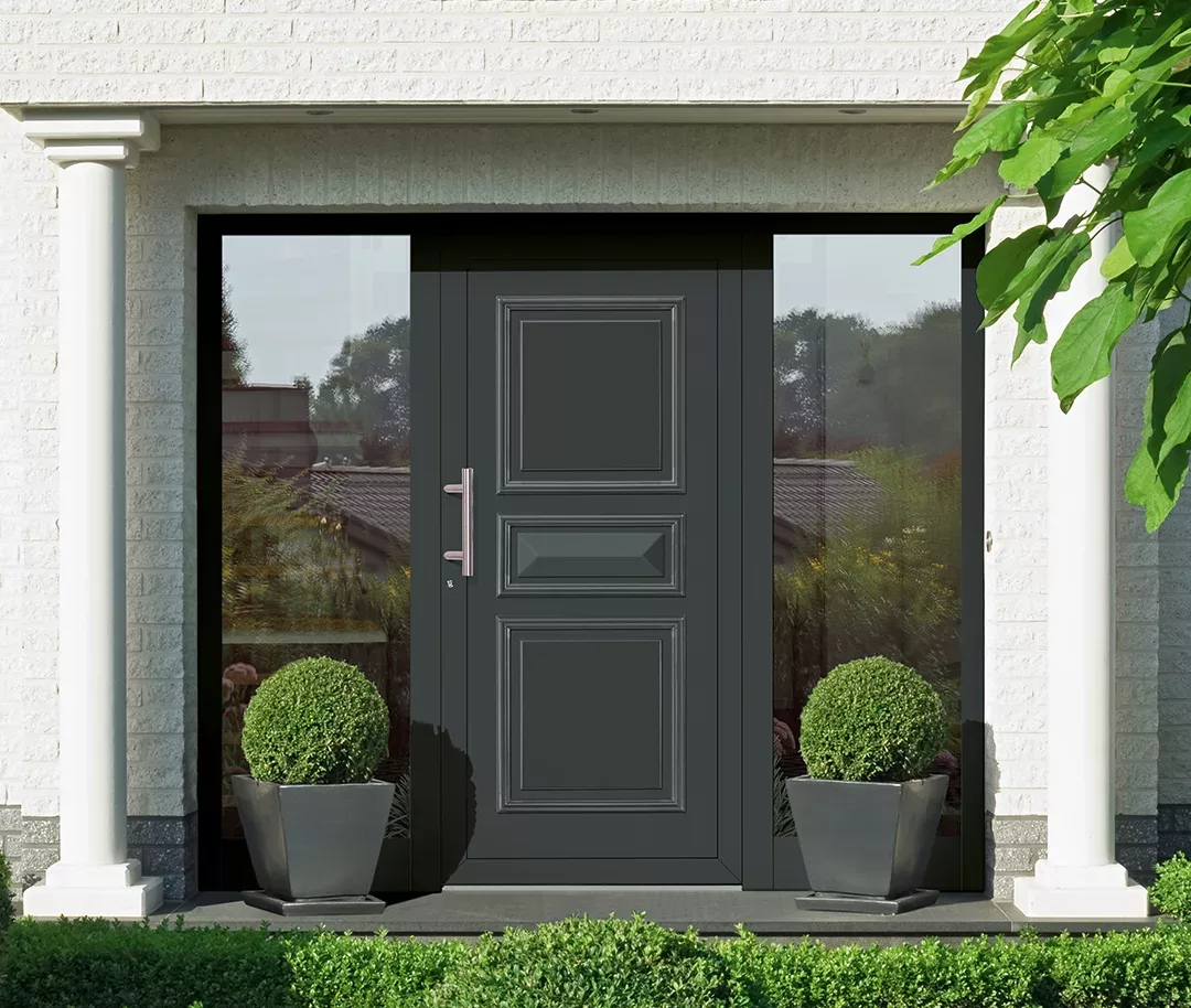 Ciemnoszare drzwi wejściowe z frezowanymi panelami i wąskimi bocznymi naświetlami, między białymi kolumnami, z dwiema kulistymi roślinami w kwadratowych donicach na tle jasnej elewacji z cegły.