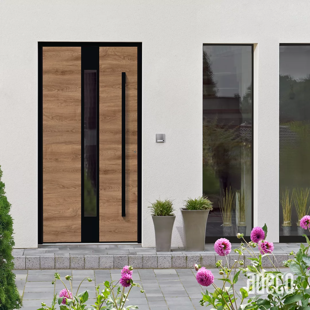 Nowoczesne wejście domu z drewnopodobnymi drzwiami z pionowym czarnym przeszkleniem i długim pochwytem, biała elewacja, wąskie okno boczne oraz donice i różowe kwiaty na pierwszym planie.