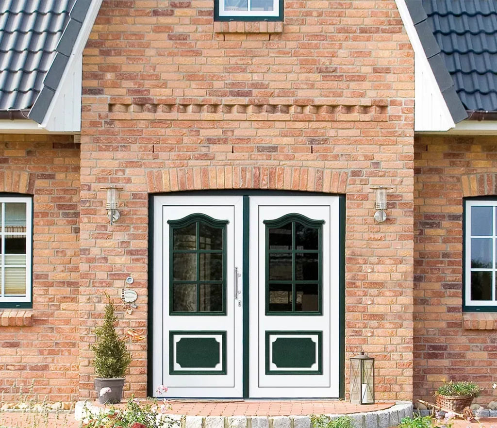 Wejście do ceglanego domu: białe dwuskrzydłowe drzwi z zielonymi panelami i przeszkleniami pod ceglanym łukiem, po bokach kinkiety, a na ganku donica i latarnia.
