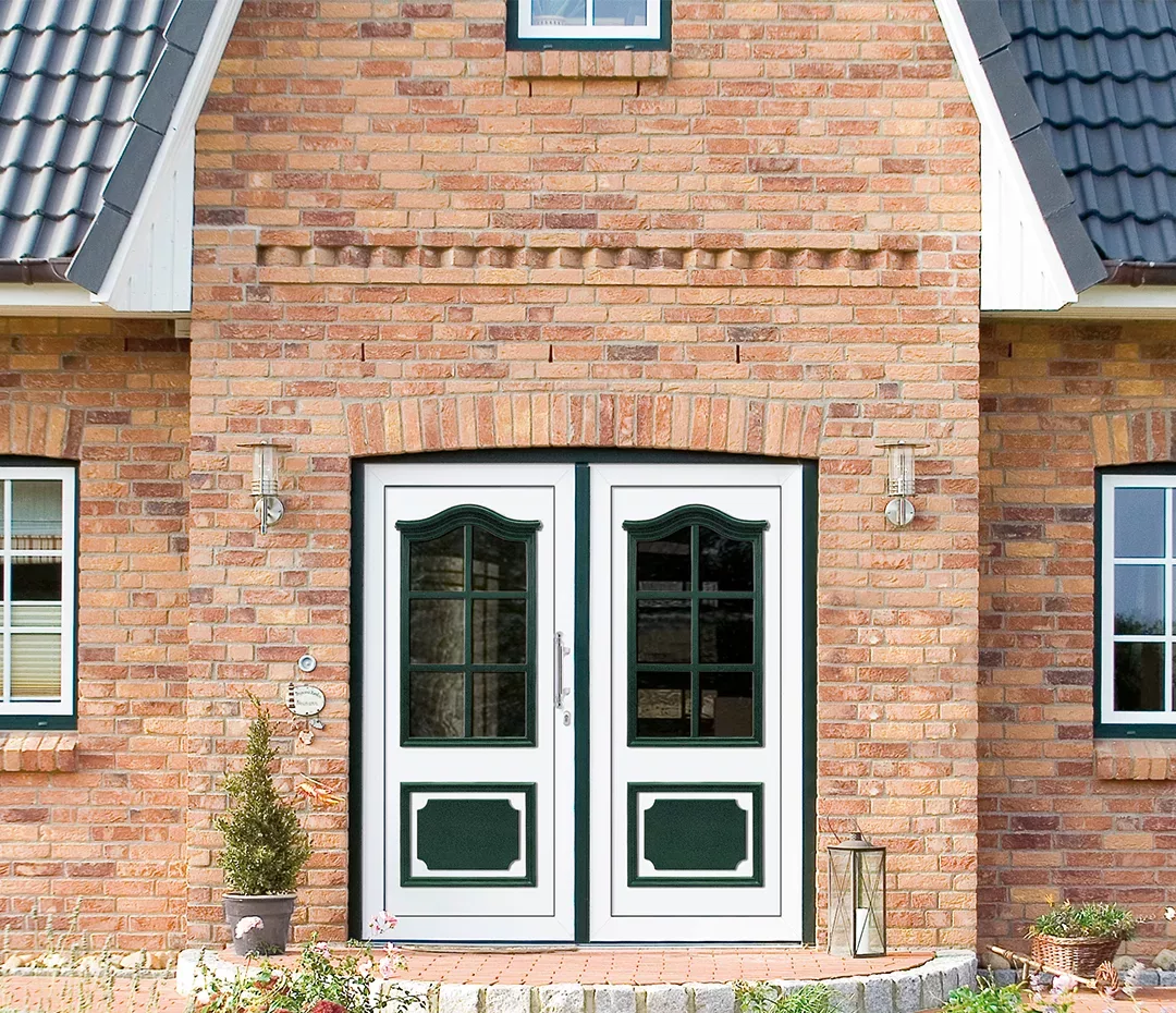 Wejście do ceglanego domu: białe dwuskrzydłowe drzwi z zielonymi panelami i przeszkleniami pod ceglanym łukiem, po bokach kinkiety, a na ganku donica i latarnia.