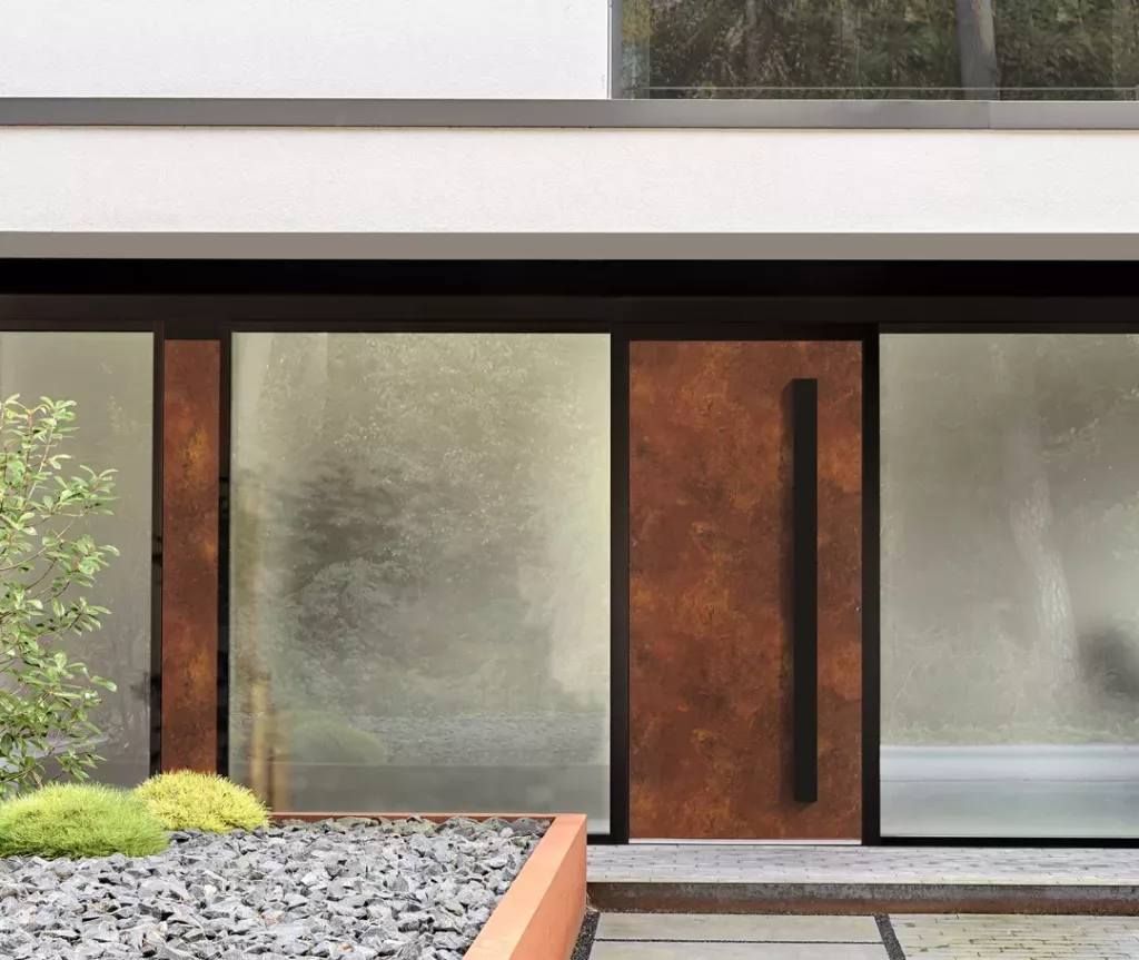 Nowoczesne wejście do domu z dużymi matowymi przeszkleniami, drzwiami ze stali corten z długim pionowym uchwytem oraz minimalistyczną rabatą żwirową z niskimi krzewami.