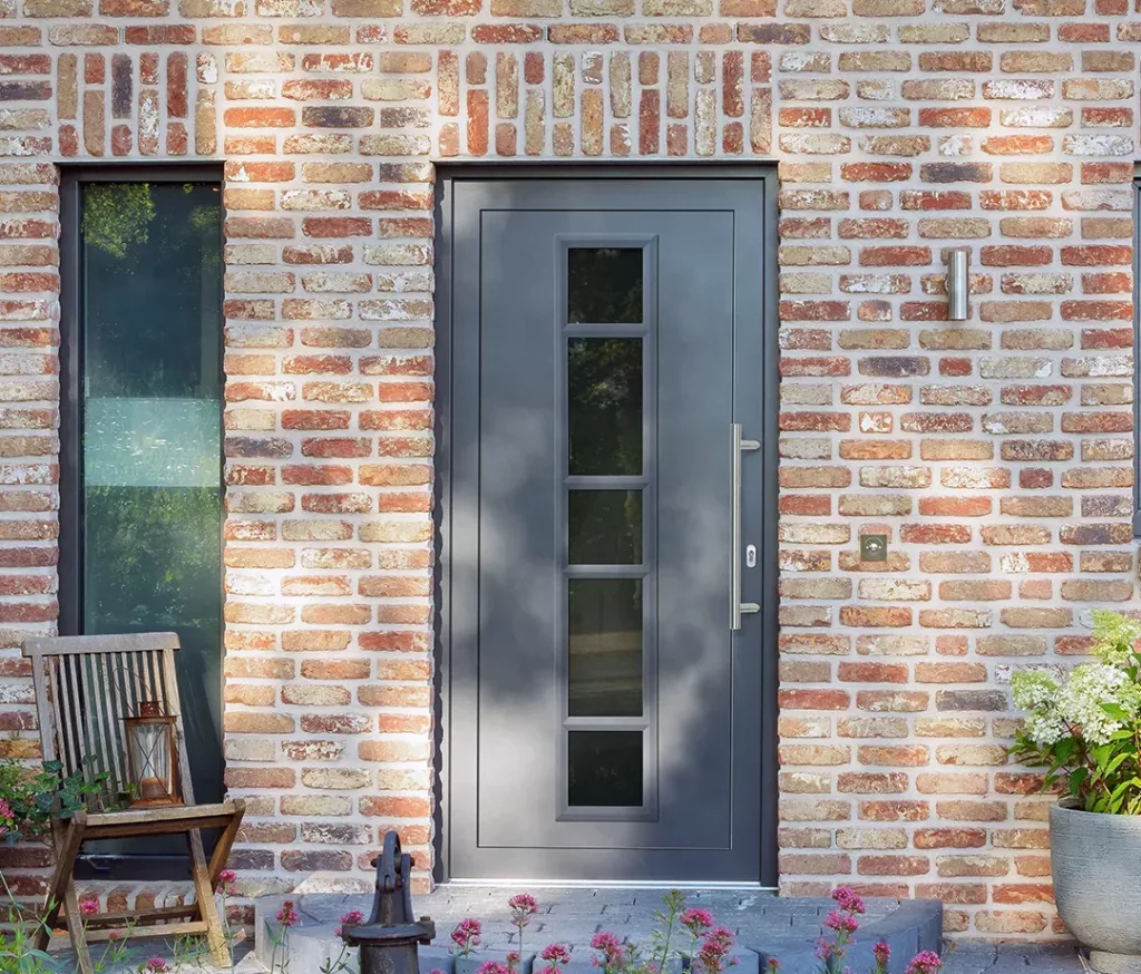 Ciemnoszare drzwi wejściowe z pionowym pasem czterech szyb w ceglanej elewacji, obok wąskie okno, kinkiet i dzwonek, przy progu drewniane krzesło z lampionem oraz donice z roślinami.