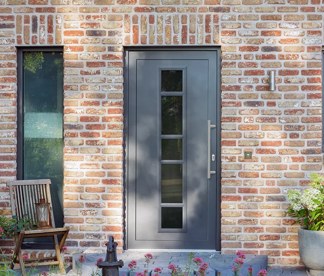 Ciemnoszare drzwi wejściowe z pionowym pasem czterech szyb w ceglanej elewacji, obok wąskie okno, kinkiet i dzwonek, przy progu drewniane krzesło z lampionem oraz donice z roślinami.