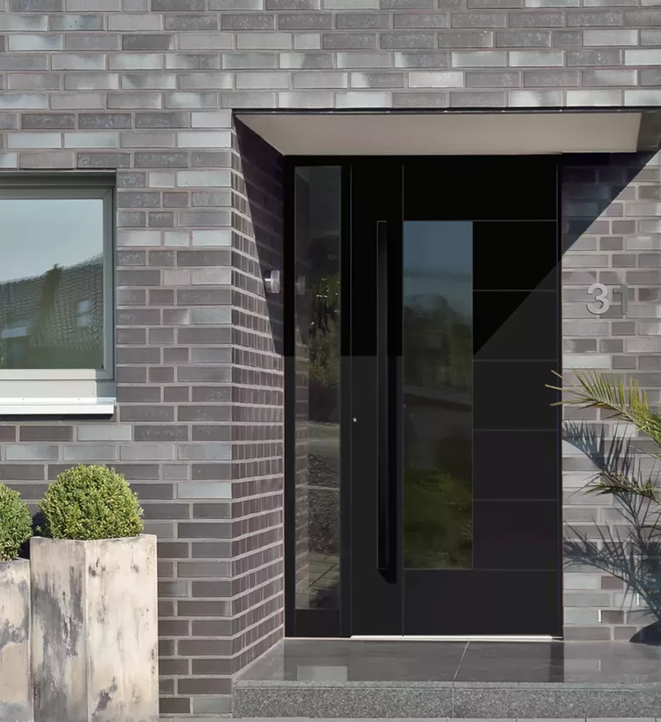 Nowoczesne czarne drzwi wejściowe z pionowym przeszkleniem i długim uchwytem w elewacji z szarej cegły, obok okna i numeru 31, z roślinami w donicach przy progu.