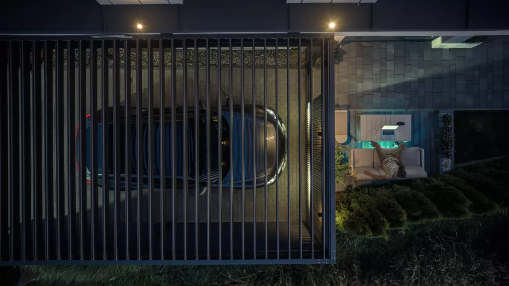 Widok z góry na zaparkowany samochód pod nowoczesną wiatą z lamelami obok tarasu, gdzie mężczyzna odpoczywa na sofie przy stoliku z laptopem, w wieczornym oświetleniu i otoczeniu zieleni.