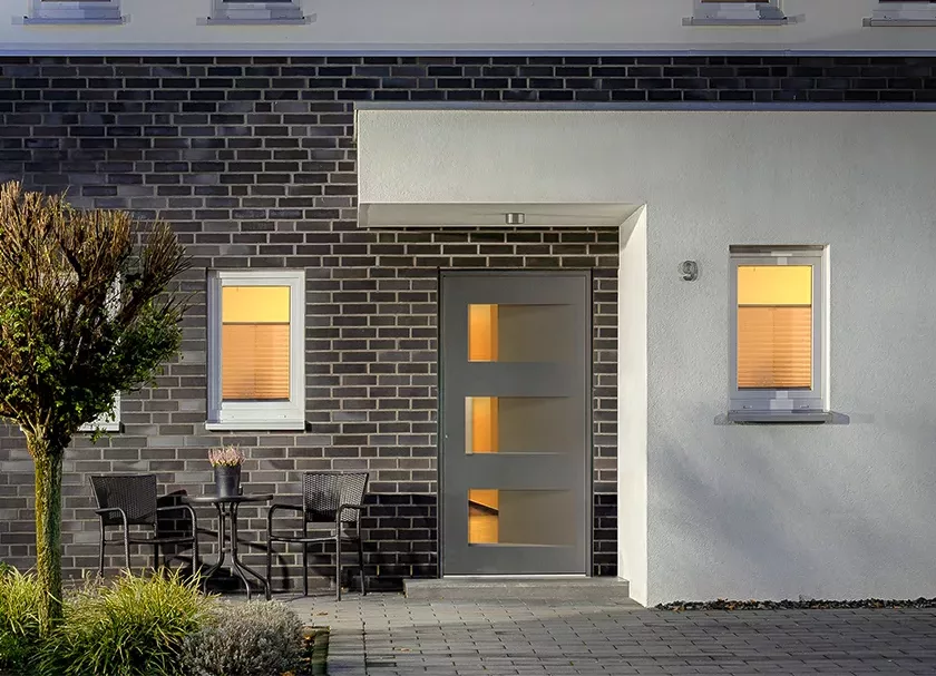 Nowoczesne wejście do domu: szare drzwi z trzema poziomymi przeszkleniami, elewacja z ciemnej cegły i białego tynku, dwa podświetlone okna oraz małe patio z dwoma krzesłami i stolikiem z rośliną.