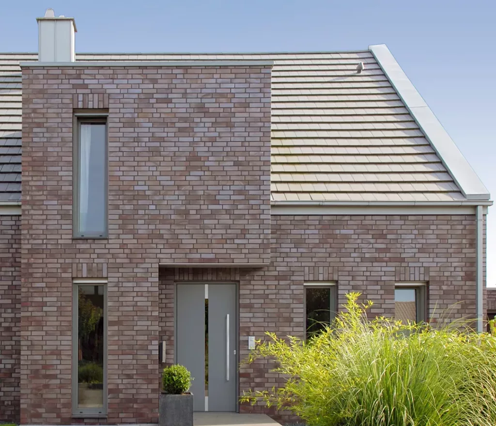 Front nowoczesnego domu z cegły w szaro‑brązowym odcieniu, z wąskimi pionowymi oknami, centralnymi szarymi drzwiami oraz dachem z jasnoszarych dachówek, otoczony prostą zielenią z trawą ozdobną.