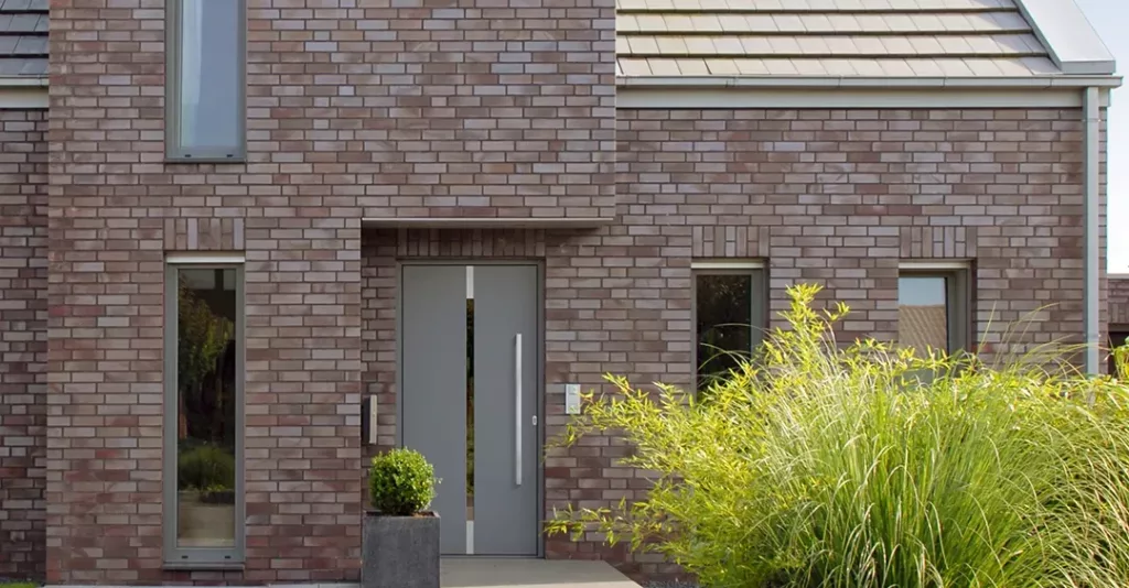 Front nowoczesnego domu z cegły w szaro‑brązowym odcieniu, z wąskimi pionowymi oknami, centralnymi szarymi drzwiami oraz dachem z jasnoszarych dachówek, otoczony prostą zielenią z trawą ozdobną.