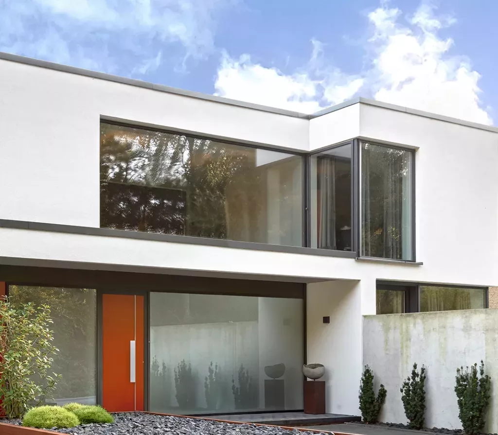 Nowoczesny, minimalistyczny dom z białą elewacją, dużymi narożnymi przeszkleniami i płaskim dachem, z pomarańczowymi drzwiami wejściowymi oraz ogrodem z kamykami i niskimi krzewami na tle błękitnego nieba.