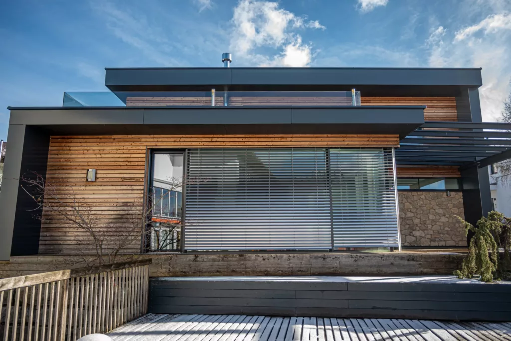 Nowoczesna fasada domu z drewnianą okładziną, płaskim dachem i dużym oknem przesłoniętym zewnętrznymi żaluzjami, z tarasem z desek pod jasnym niebem.