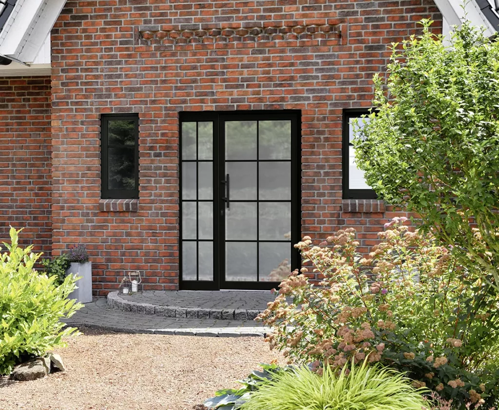 Front domu z czerwonej cegły z czarnymi drzwiami przeszklonymi matowym szkłem drzwiami przedzielone czarnymi szprosami z dostawką boczną, dwiema wąskimi oknami i kamiennym podestem, otoczony zadbanym ogrodem ze krzewami.