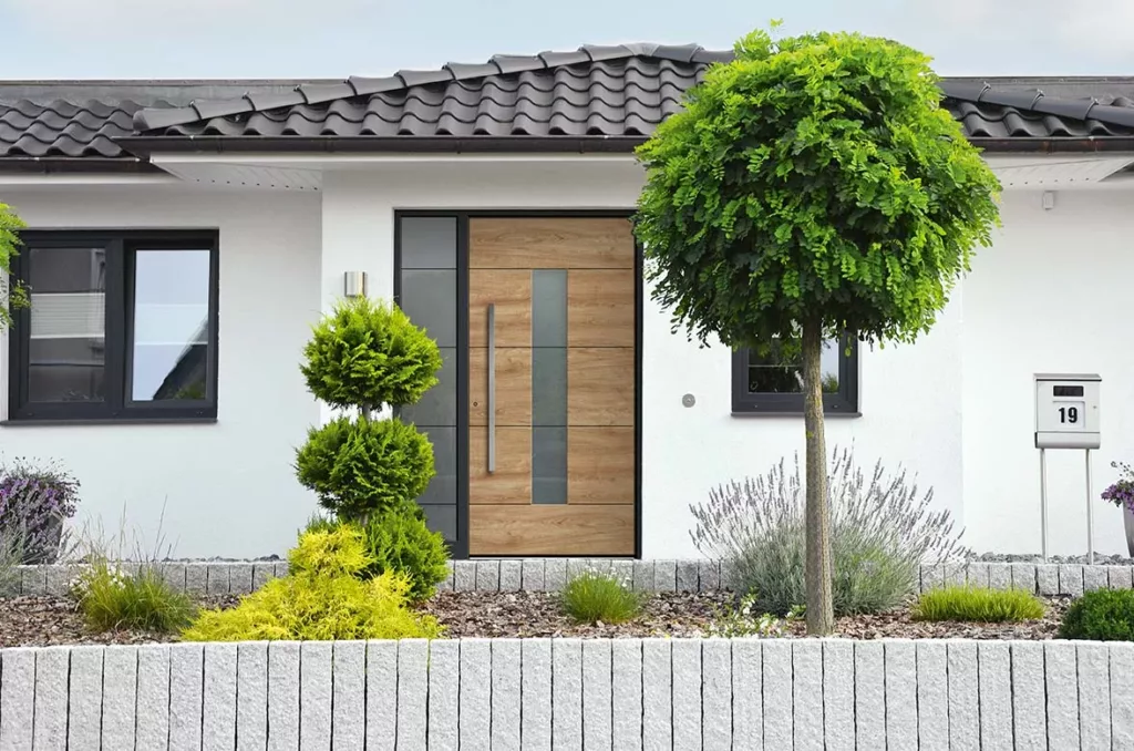 Nowoczesne wejście do domu z drzwiami zewnętrznymi z drewnopodobnym panelem drzwiowym z pionowym pasem szkła i z dostawką boczną ze szkła po lewej stronie, białą elewacją i oknami z profilami okiennymi o kolorze antracytu, otoczone minimalistycznym ogrodem z formowanymi krzewami i drzewkiem; po prawej skrzynka pocztowa z numerem 19.