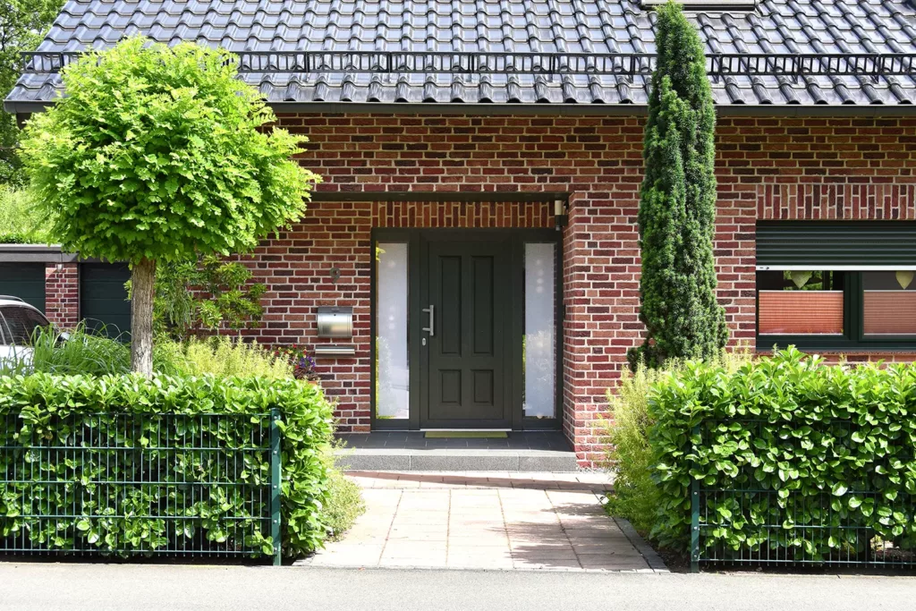 Wejście do domu z czerwonej cegły z ciemnymi drzwiami i dostawką boczną z mlecznej szyby, do którego prowadzi brukowana ścieżka, otoczone żywopłotem, drzewkiem o kulistej koronie i smukłym iglakiem.